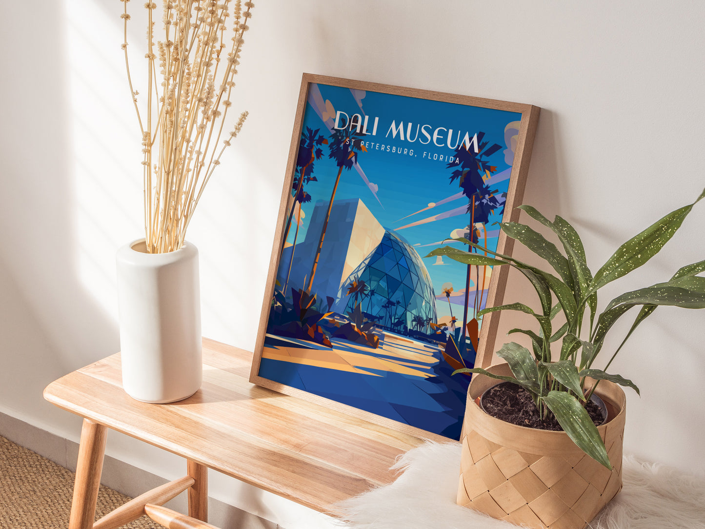 A framed Dali Museum poster with vibrant colors sits on a wooden bench next to a white vase with dried flowers and a potted plant, creating a stylish decor setup.