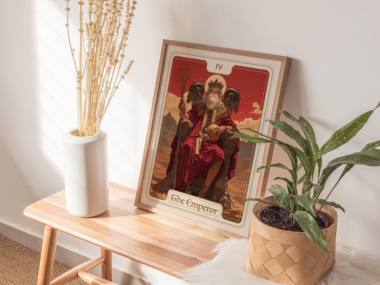 A framed Emperor Tarot Card poster sits on a wooden table beside a white vase with dried flowers and a potted green plant.
