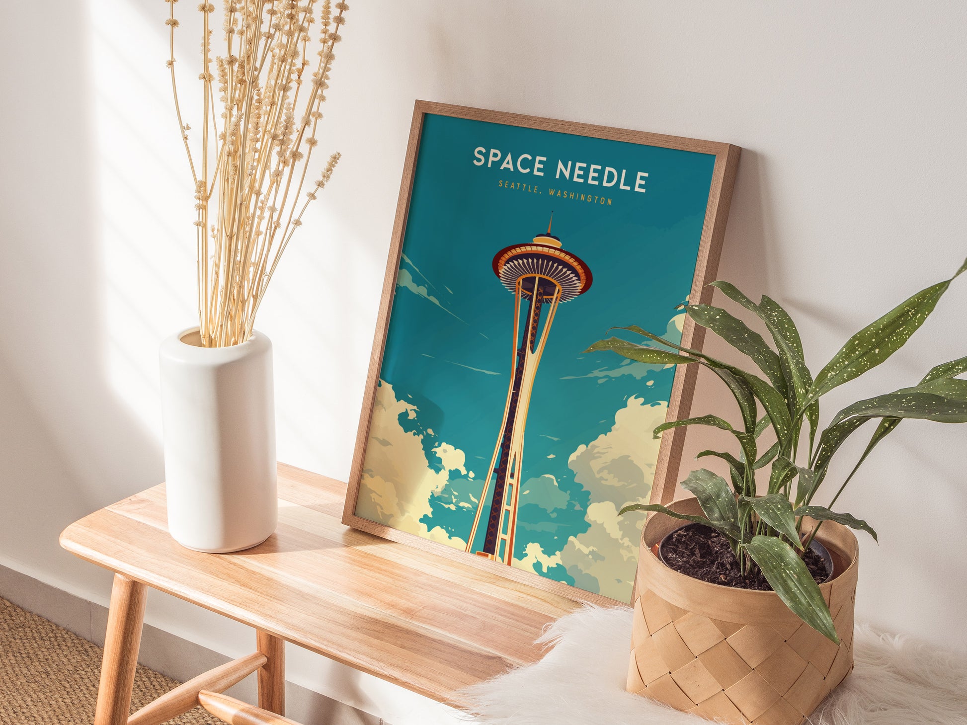 A framed Space Needle poster leans against a wall on a wooden bench, flanked by a white vase with dried flowers and a potted green plant.