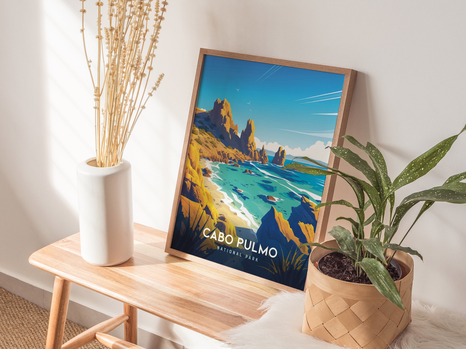 A colorful Cabo Pulmo National Park poster leans on a wooden bench beside a plant and a vase with dried stems.