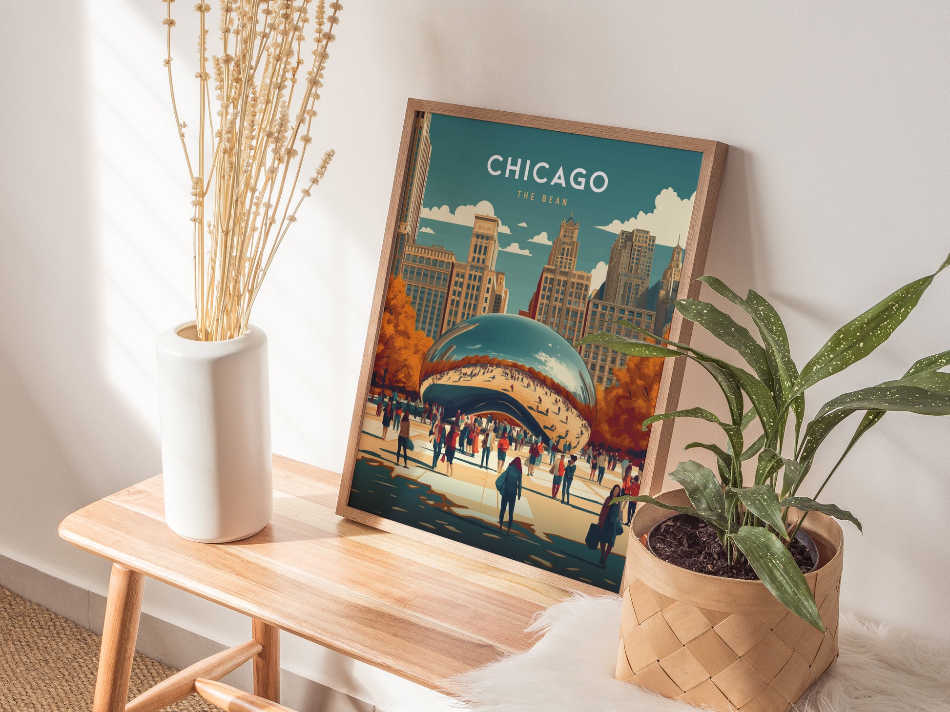 A framed poster of the Chicago Bean in Millennium Park sits on a wooden bench, accompanied by a white vase with dried flowers and a potted green plant.