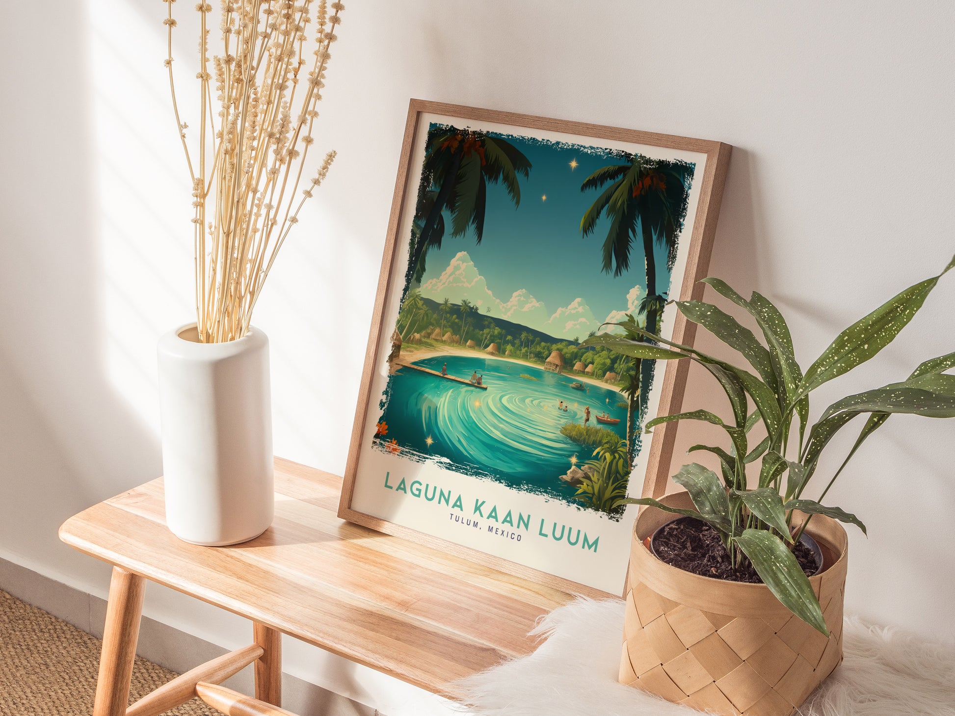 A framed Laguna Kaan Luum poster rests on a wooden bench beside a white vase with dried flowers and a potted plant, creating a serene, natural display.