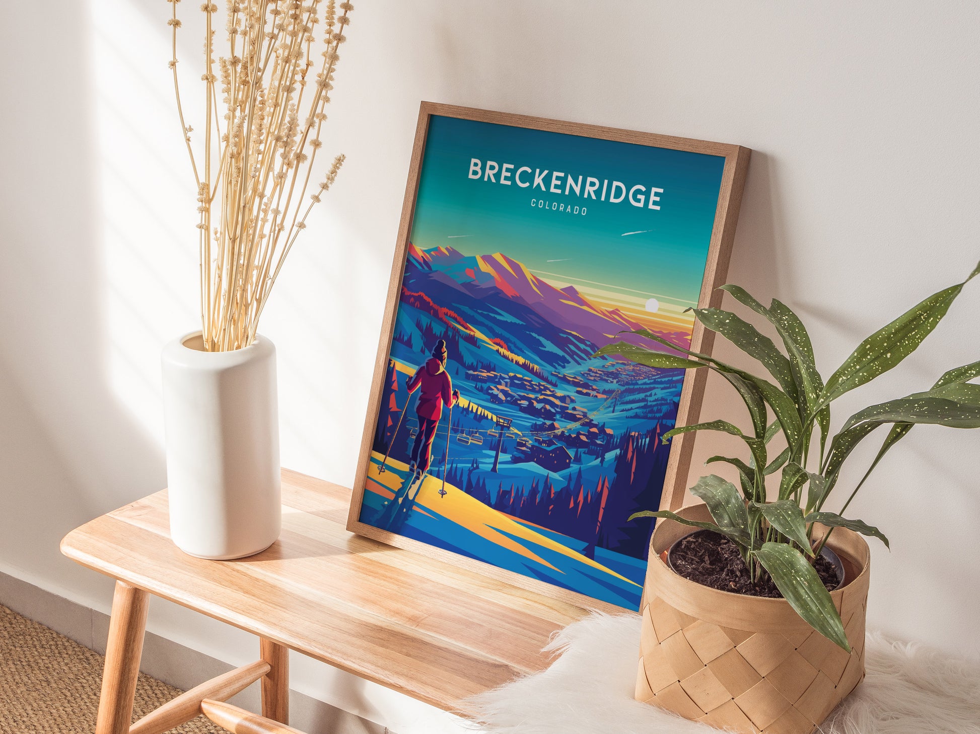 A framed Breckenridge Colorado ski poster rests on a wooden bench, accompanied by a white vase with dried flowers and a green potted plant, creating a cozy, decorative corner.