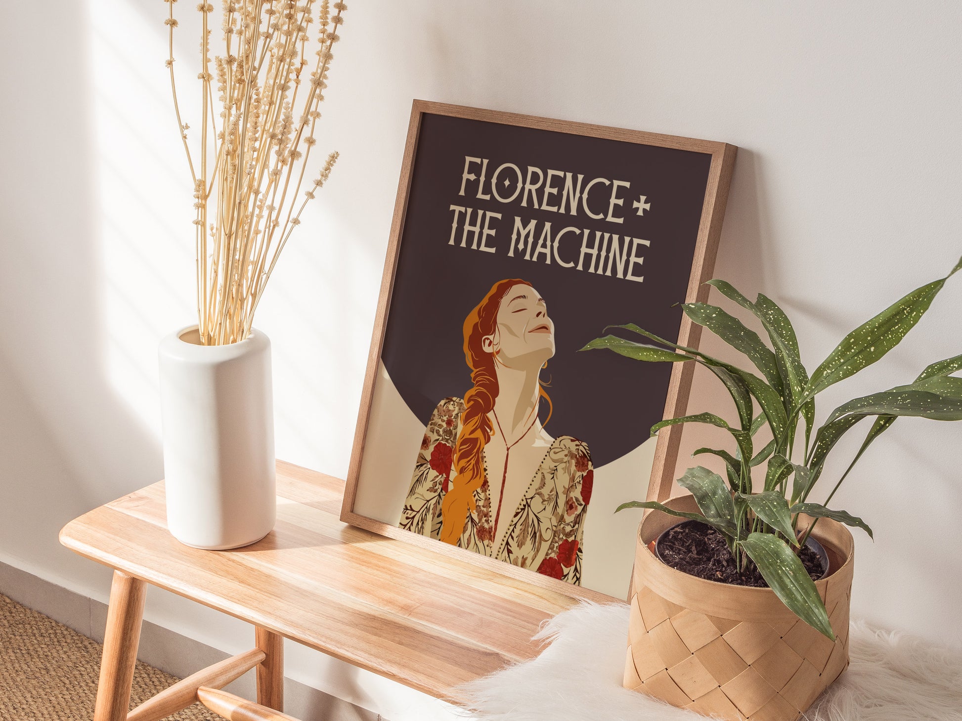 A Florence + The Machine poster rests on a wooden table between a white vase with dried plants and a woven basket with a green plant.