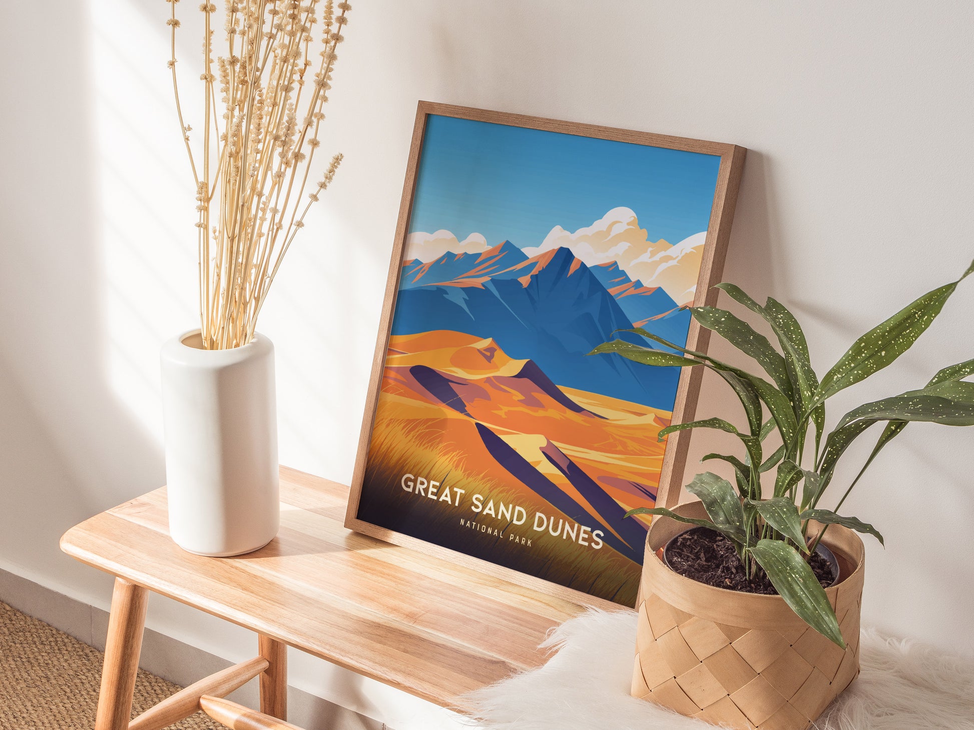 A framed Great Sand Dunes poster sits on a wooden bench beside a white vase with dried flowers and a green potted plant. Sunlight casts shadows on the wall.