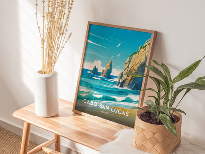 A framed Cabo San Lucas poster with vibrant ocean and cliff imagery is displayed on a wooden bench, accompanied by a white vase of dried flowers and a potted plant.