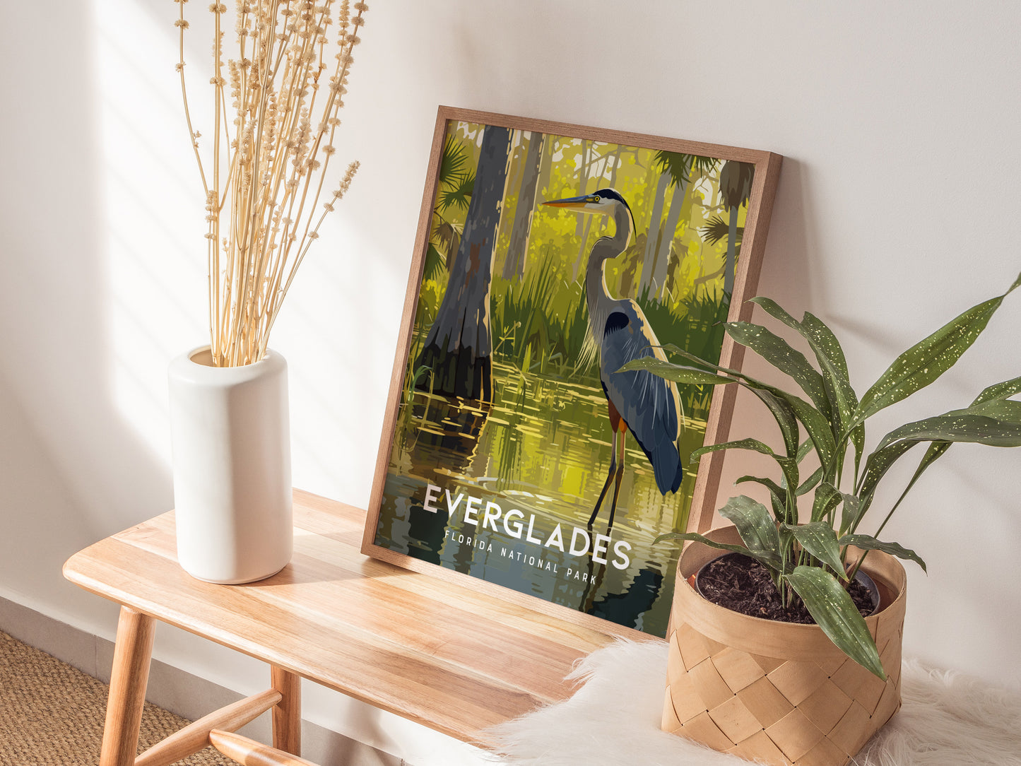 A framed Everglades poster featuring a Great Blue Heron rests on a wooden table beside a white vase with dried stems and a potted green plant.