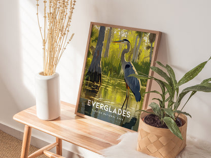 A framed Everglades poster featuring a Great Blue Heron rests on a wooden table beside a white vase with dried stems and a potted green plant.