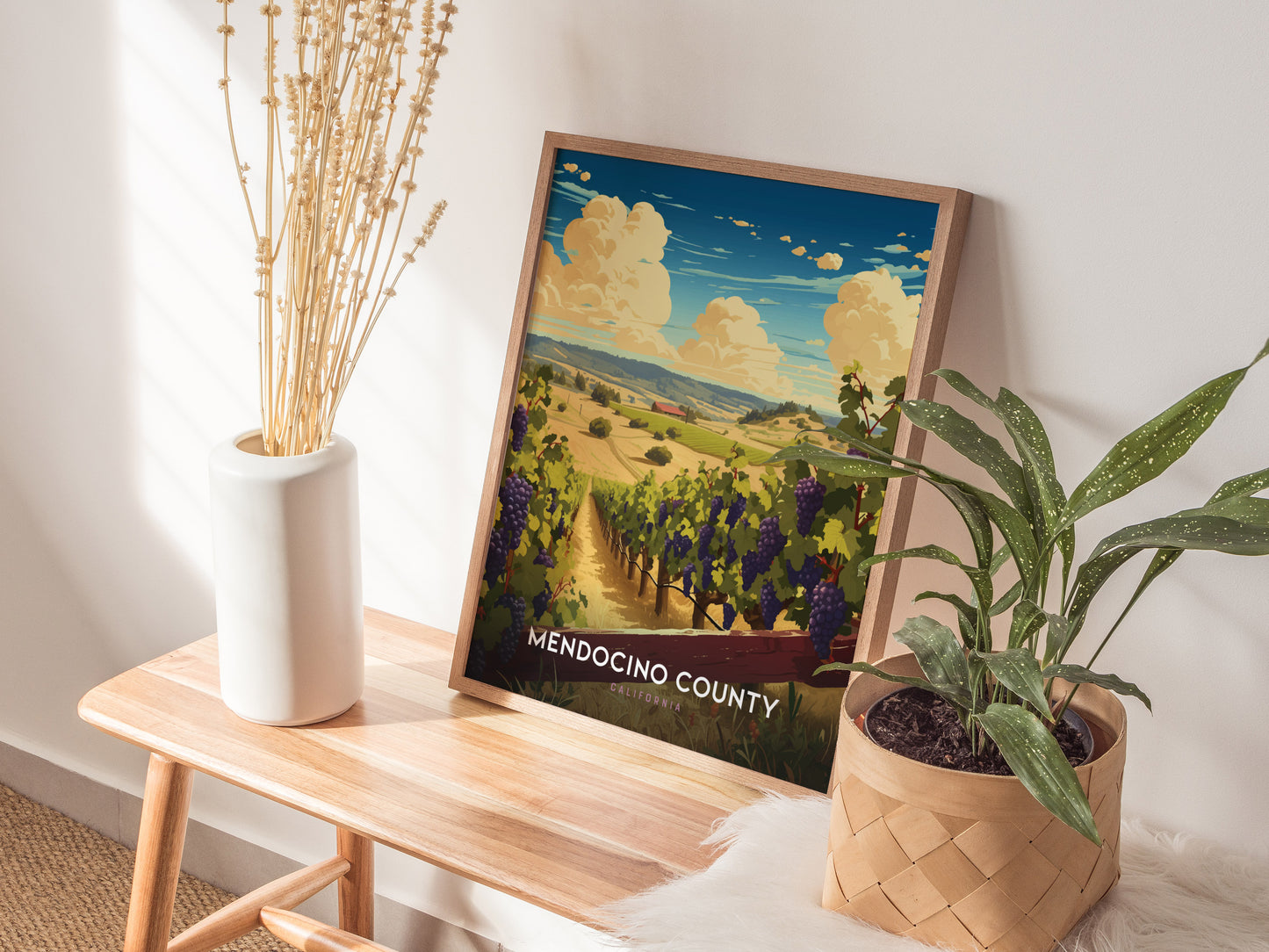 A framed Mendocino County poster depicting vineyards and rolling hills rests on a wooden bench beside a white vase with dried flowers and a potted green plant.