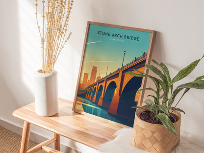 A framed Stone Arch Bridge poster rests on a wooden table beside a white vase with dried flowers and a potted green plant. Sunlight casts soft shadows on the wall.