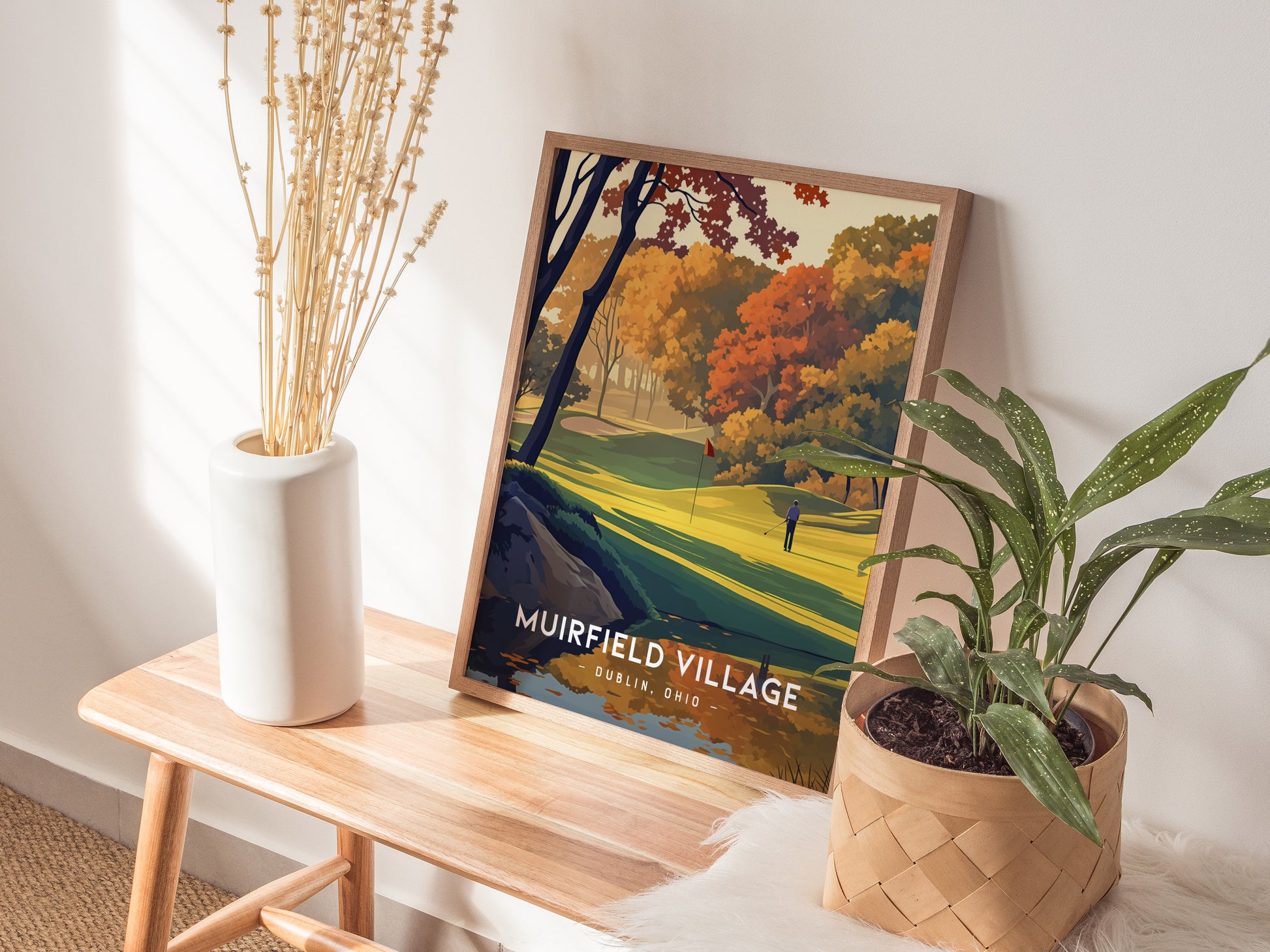 A framed Muirfield Village golf course poster leans against a wall on a wooden bench, accompanied by a white vase with dried flowers and a potted green plant.