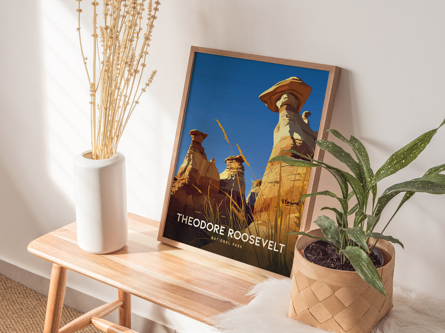 Framed poster of Theodore Roosevelt National Park featuring hoodoos rests on a wooden bench, accompanied by a white vase with dried plants and a potted green plant.