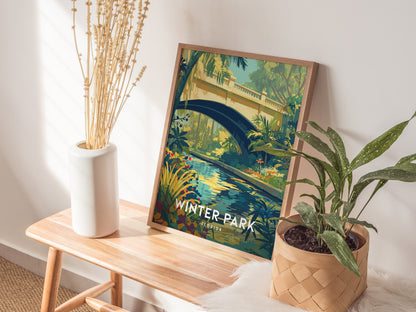 A framed poster of Winter Park, Florida, featuring a lush garden with a stream and bridge, rests on a wooden table beside a white vase with dried plants and a.