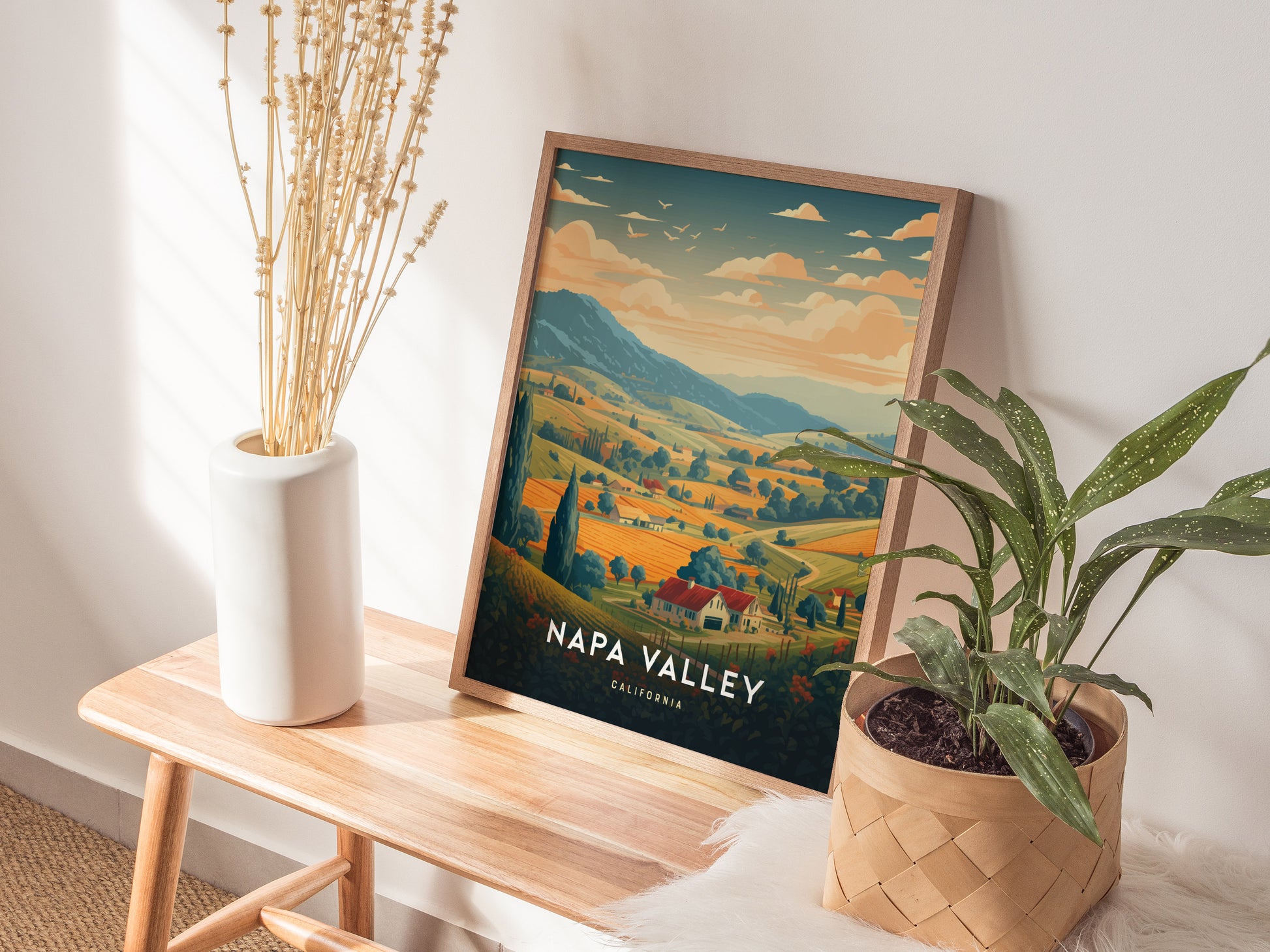 A framed Napa Valley travel poster depicting scenic vineyards and hills is displayed on a wooden bench, alongside a white vase with dried flowers and a potted plant.