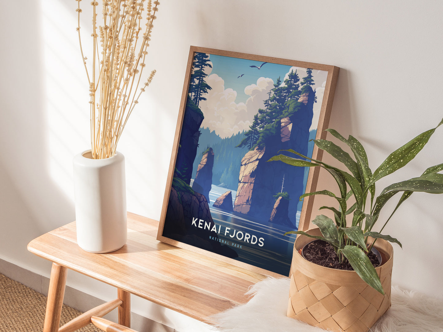 A framed Kenai Fjords poster featuring rocky cliffs and trees rests on a wooden bench beside a white vase with dried branches and a potted plant.