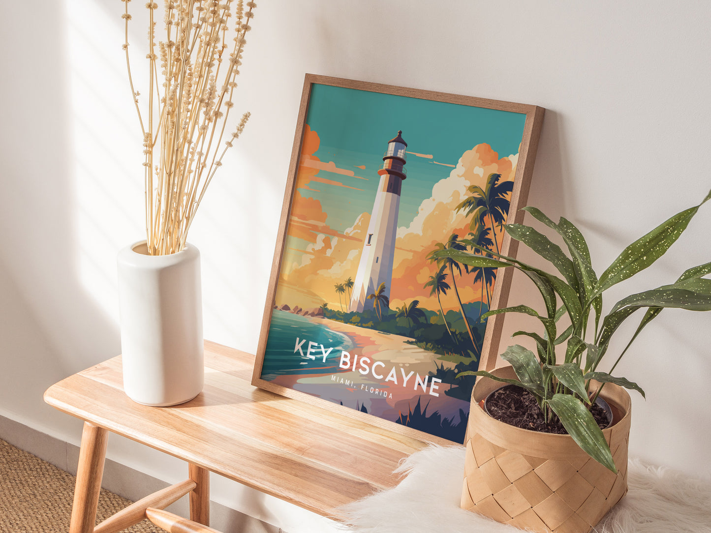 A framed Key Biscayne lighthouse poster sits on a wooden bench, surrounded by a white vase with dried plants and a potted green plant, in a sunlit room.