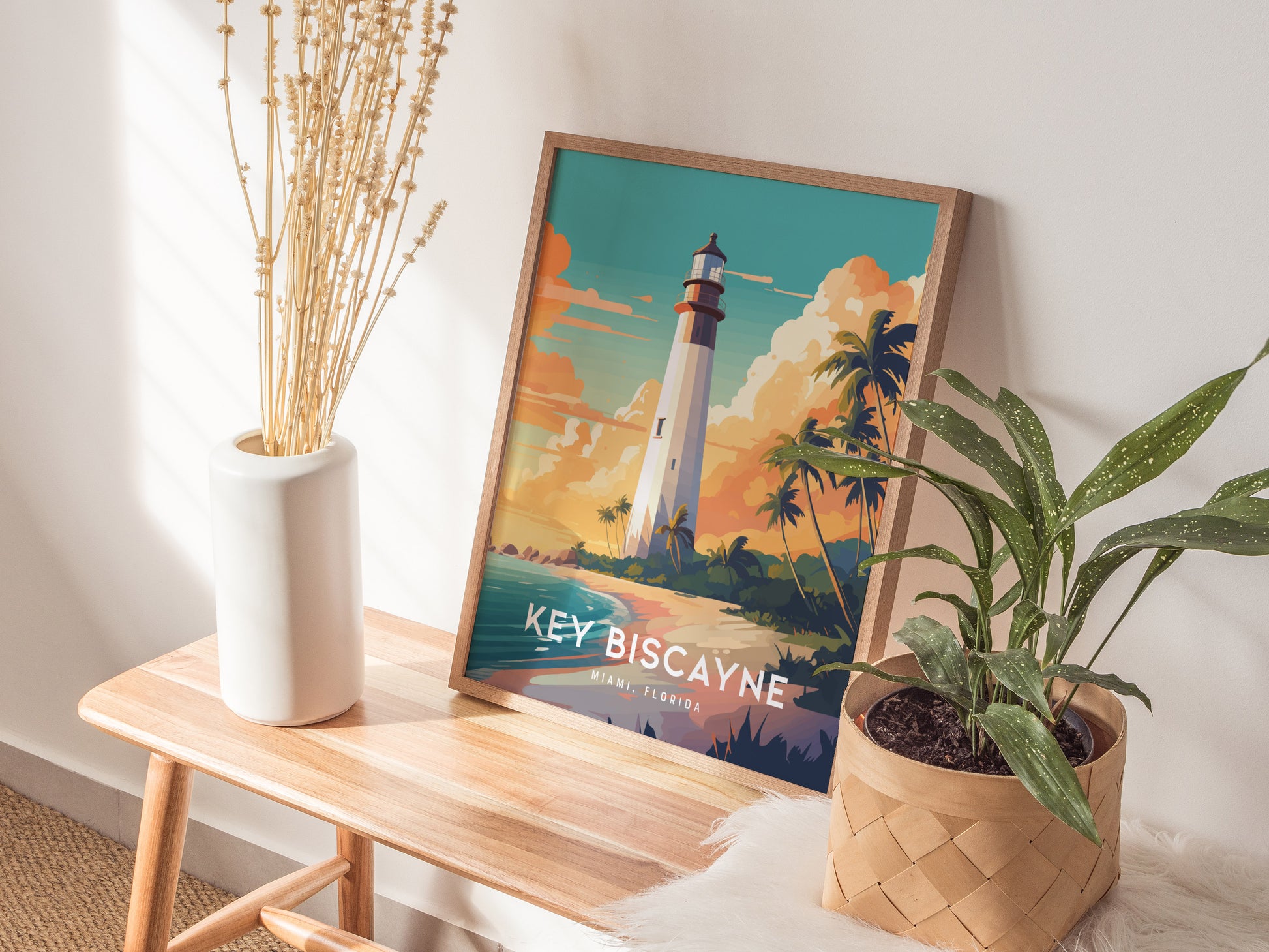 A framed Key Biscayne lighthouse poster sits on a wooden bench, surrounded by a white vase with dried plants and a potted green plant, in a sunlit room.