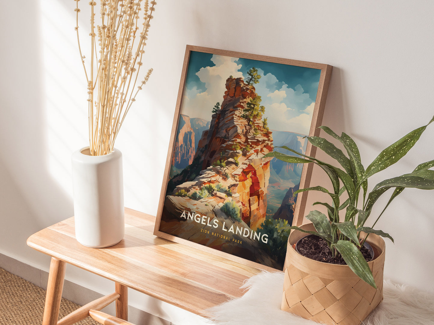 A framed Angels Landing poster rests on a wooden bench beside a white vase with dried stems and a potted plant. Sunlight casts soft shadows on the wall.