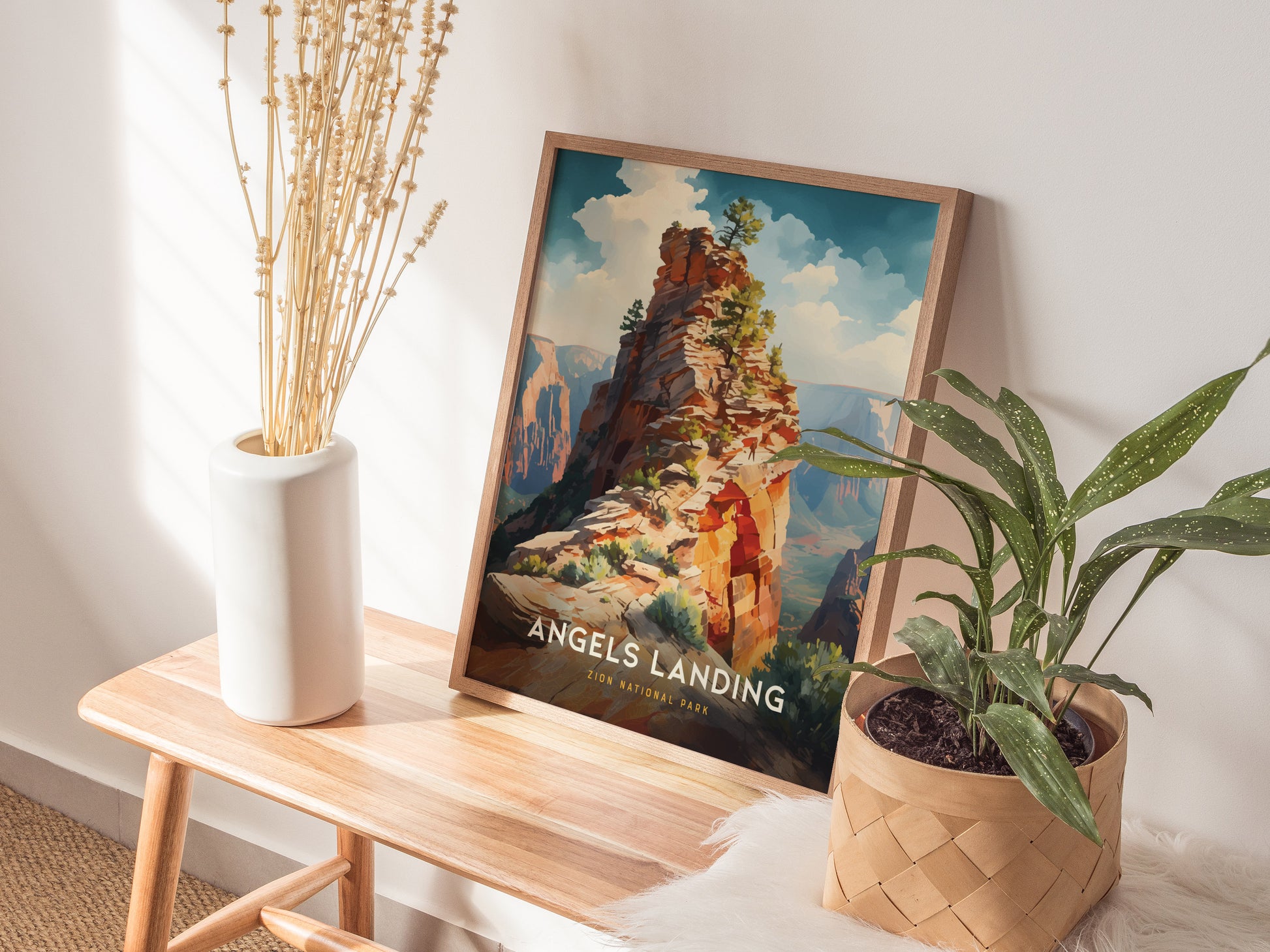 A framed Angels Landing poster rests on a wooden bench beside a white vase with dried stems and a potted plant. Sunlight casts soft shadows on the wall.