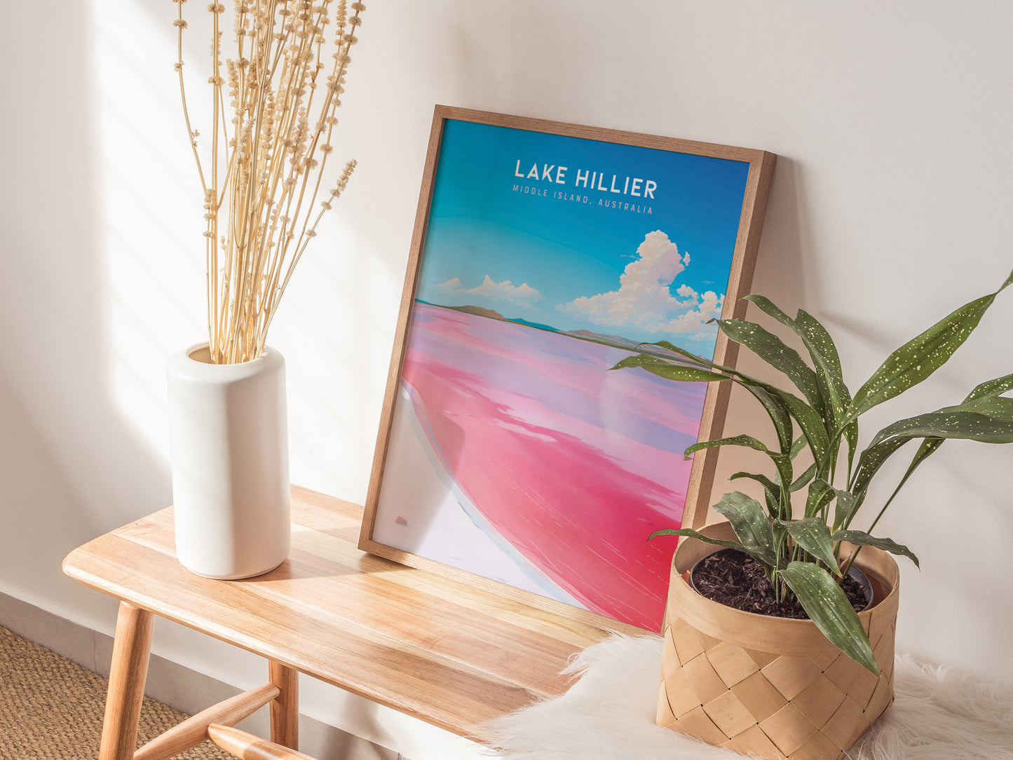 A framed Lake Hillier poster with vivid pink water and blue sky leans against a wall on a wooden bench, next to a white vase with dried flowers and a.
