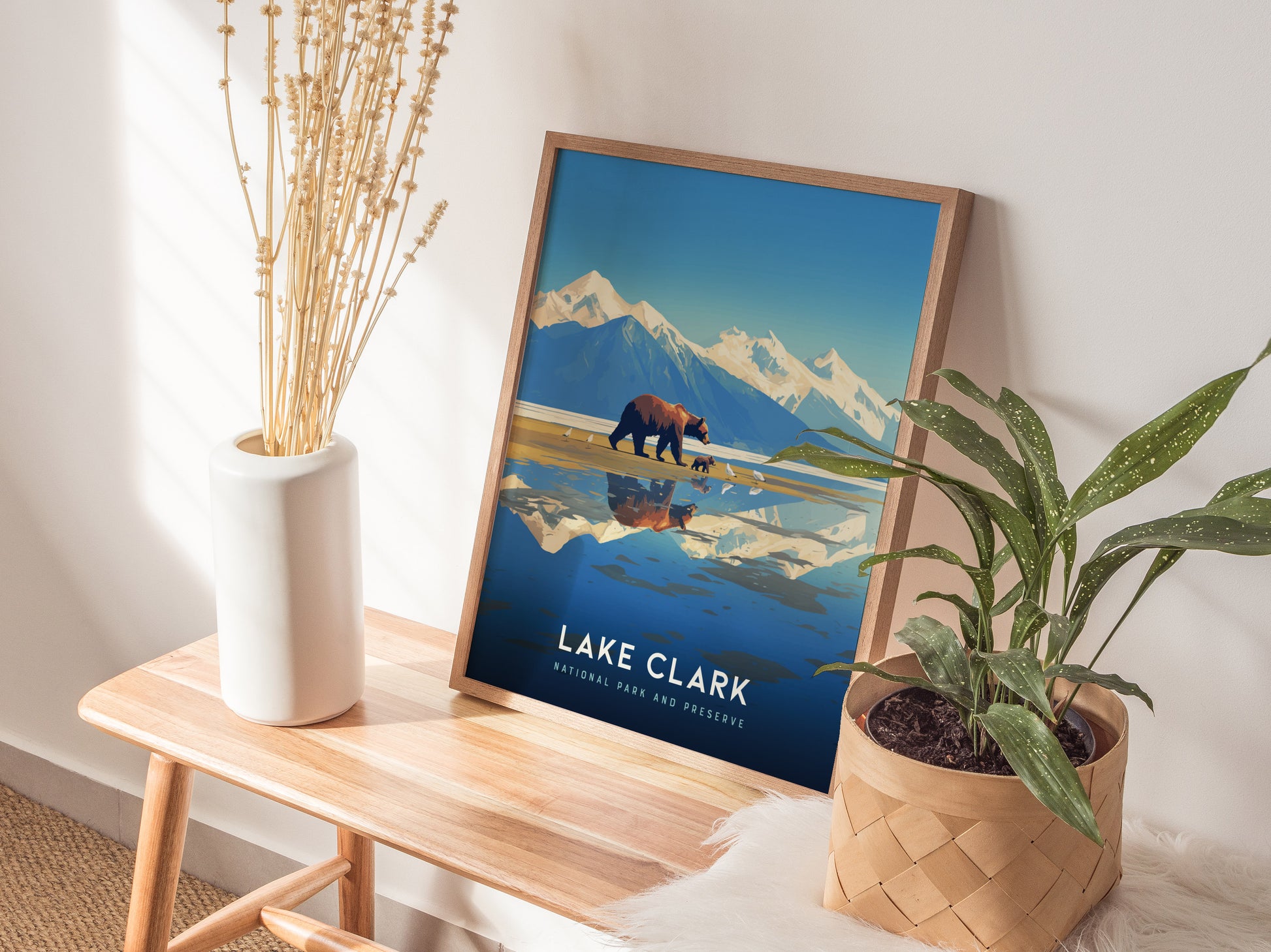 A framed Lake Clark National Park poster with mountains and a bear on a wooden bench, surrounded by a white vase with dried flowers and a potted green plant.