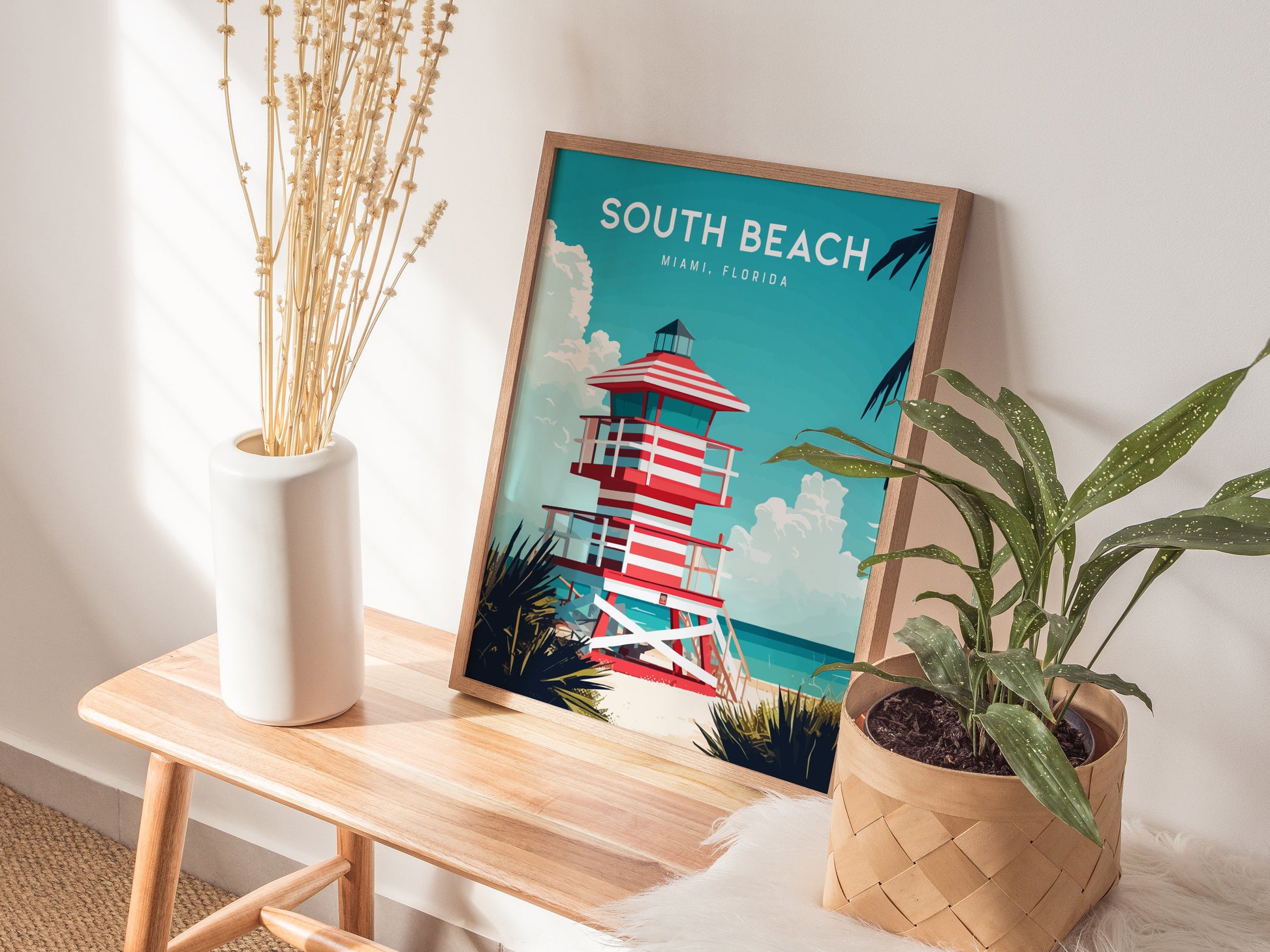 A framed South Beach Miami poster featuring a striped lifeguard tower is propped on a wooden bench next to a white vase with dried flowers and a potted plant. Sunlight casts gentle shadows across the scene.