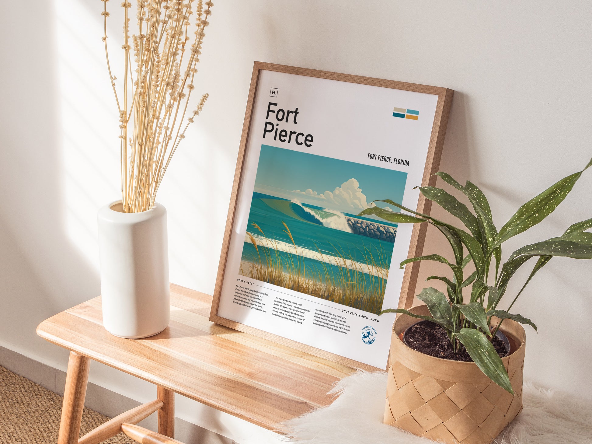 A framed Fort Pierce poster featuring a coastal scene with blue skies and ocean waves rests on a wooden bench, accompanied by a white vase with dried flowers and a.