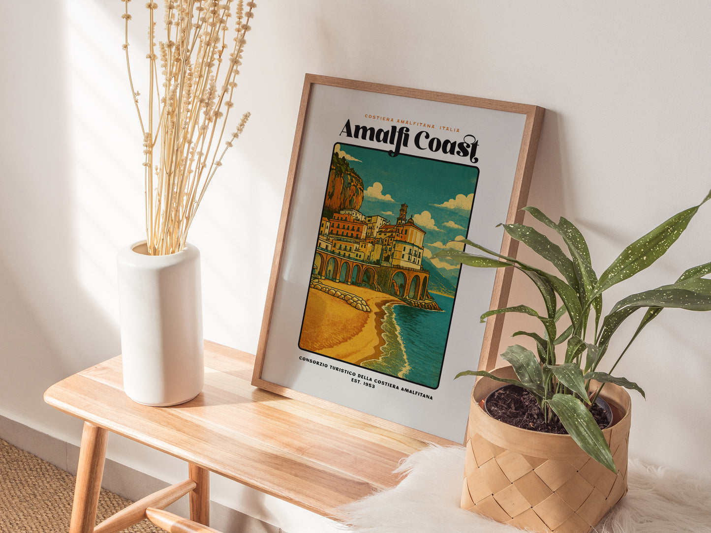 Framed Amalfi Coast poster on a wooden side table with plants and a vase.