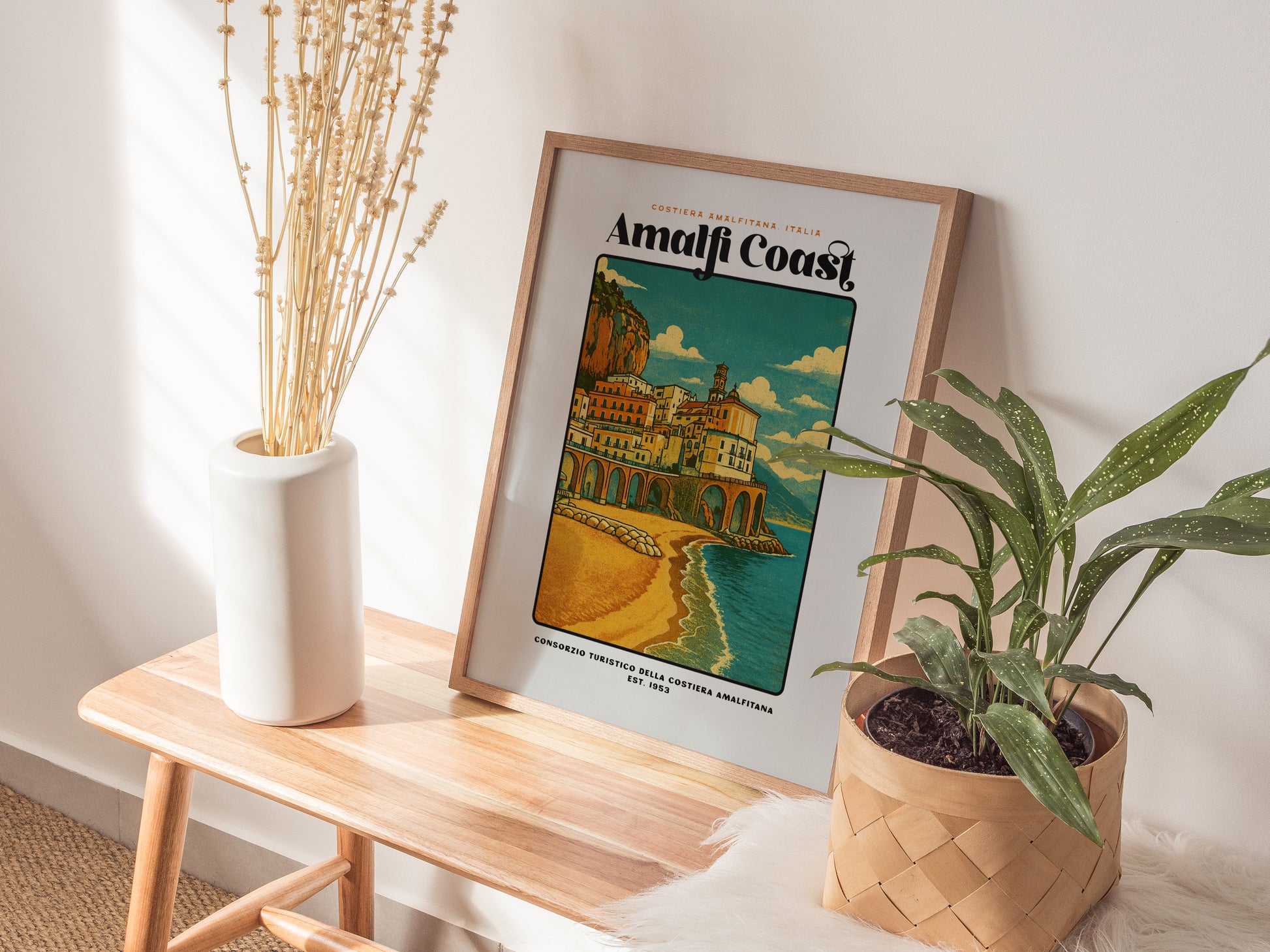 Framed Amalfi Coast poster on a wooden side table with plants and a vase.