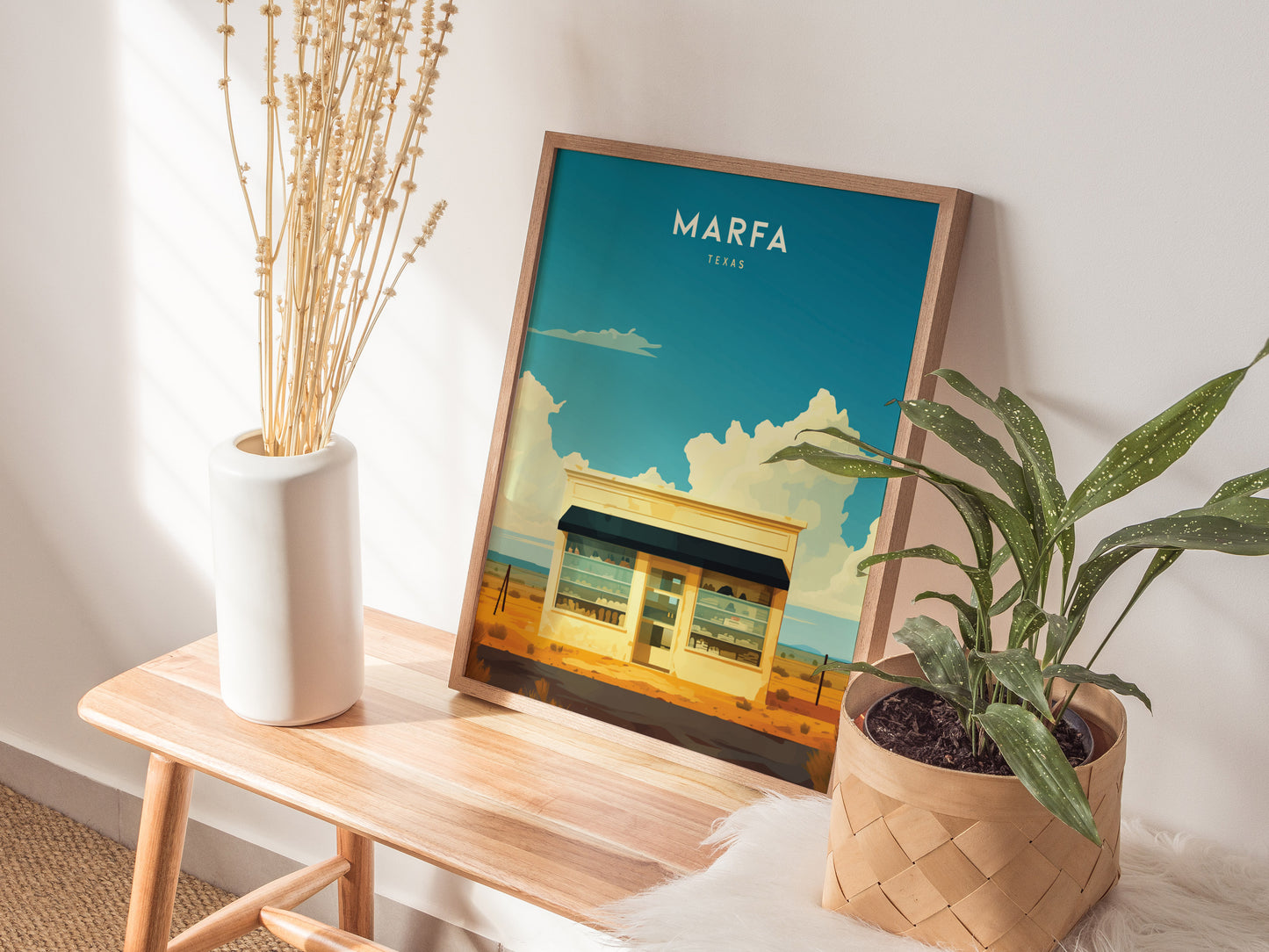 A framed Marfa Texas poster with a desert scene rests on a wooden bench, accompanied by a white vase with dried stems and a potted green plant.