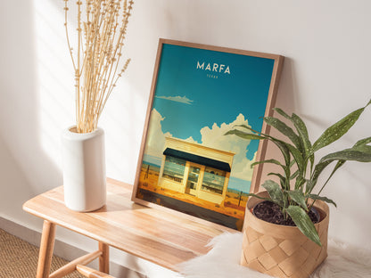 A framed Marfa Texas poster with a desert scene rests on a wooden bench, accompanied by a white vase with dried stems and a potted green plant.