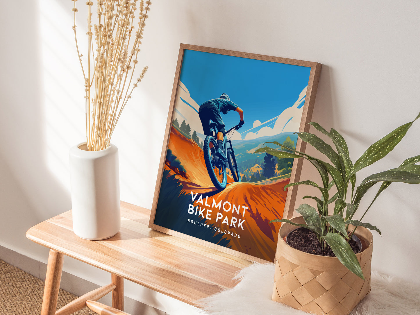 A framed Valmont Bike Park poster rests on a wooden bench beside a white vase with dried flowers and a potted plant, capturing a mountain biker against a vibrant backdrop.