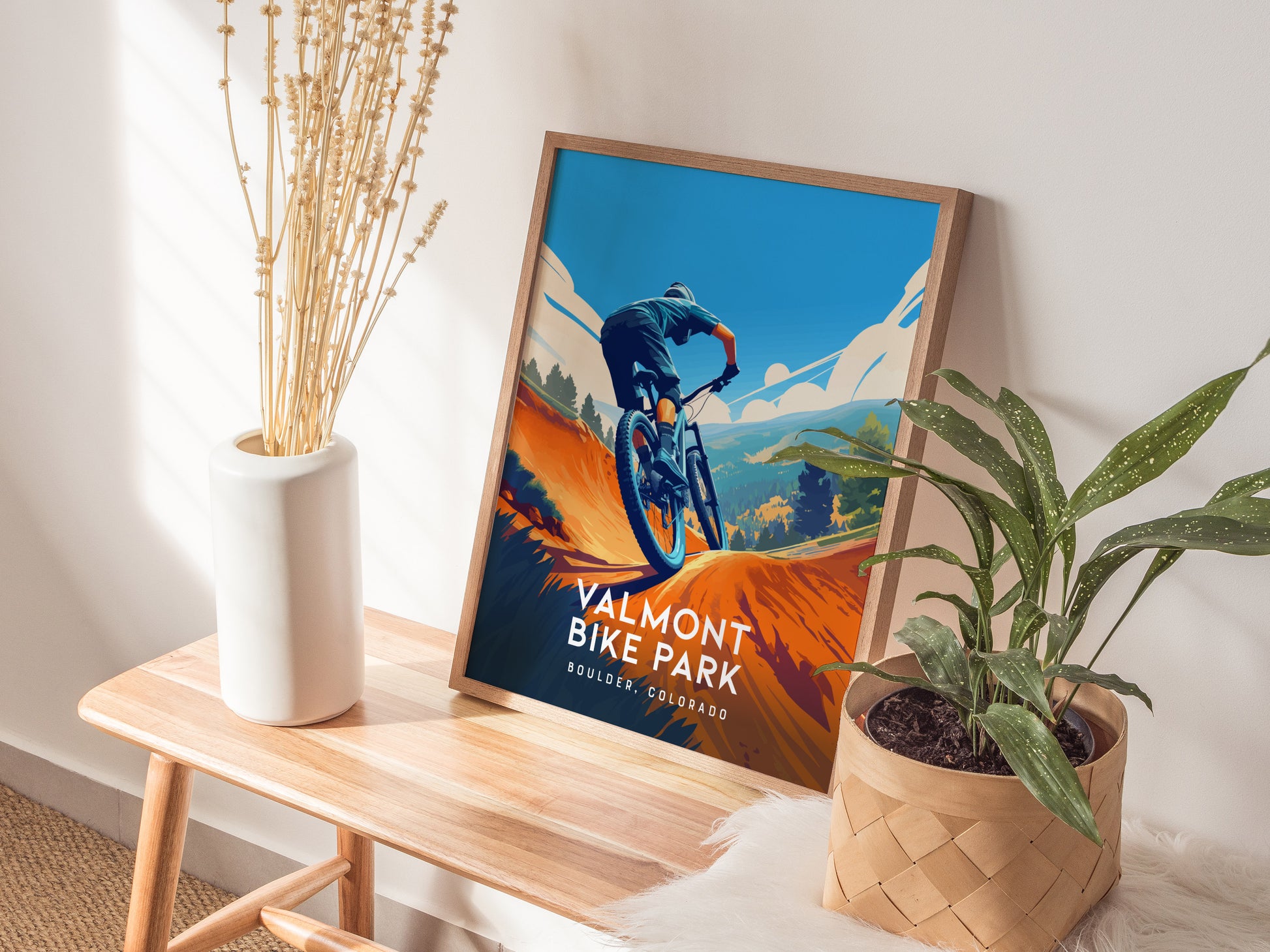 A framed Valmont Bike Park poster rests on a wooden bench beside a white vase with dried flowers and a potted plant, capturing a mountain biker against a vibrant backdrop.