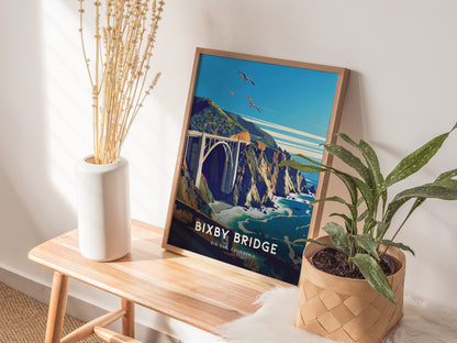 A framed poster of Bixby Bridge in Big Sur is displayed on a wooden bench, surrounded by a white vase with dried flowers and a potted green plant.