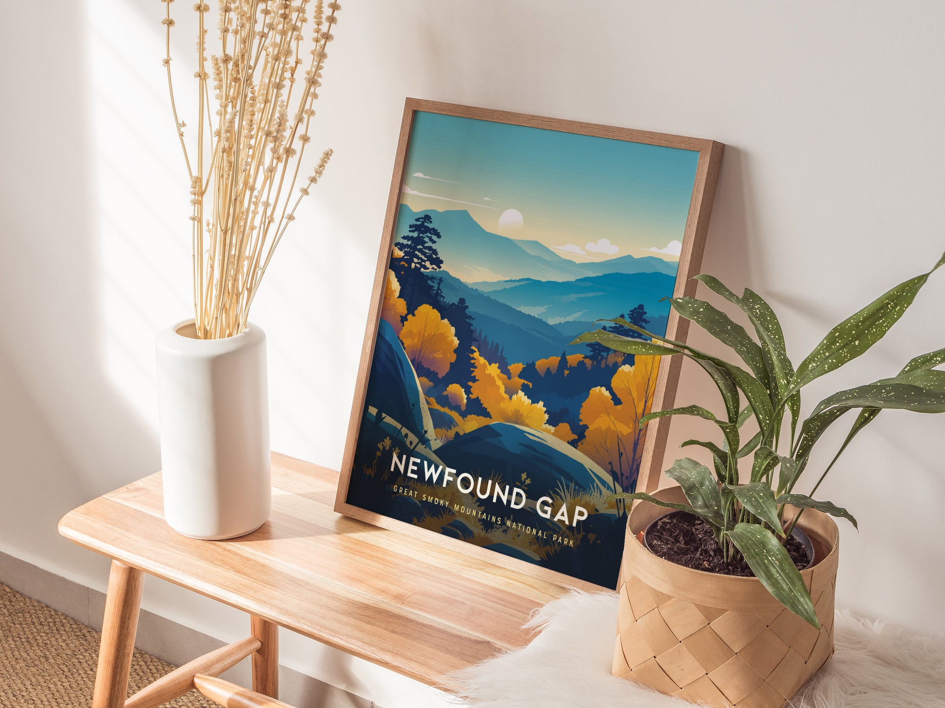 A framed Newfound Gap poster depicting blue-hued mountains and a sunlit sky rests on a wooden bench. It is surrounded by a white vase with dried stems and a potted green plant in a woven basket. Sunlight filters through a nearby window.