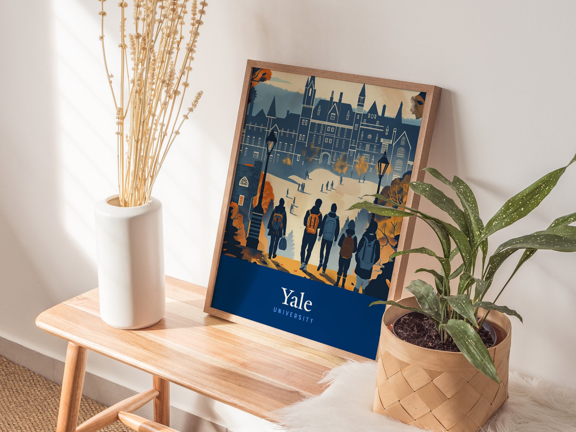 A framed poster of Yale University featuring Gothic architecture rests on a wooden bench beside a white vase with dried flowers and a potted green plant.