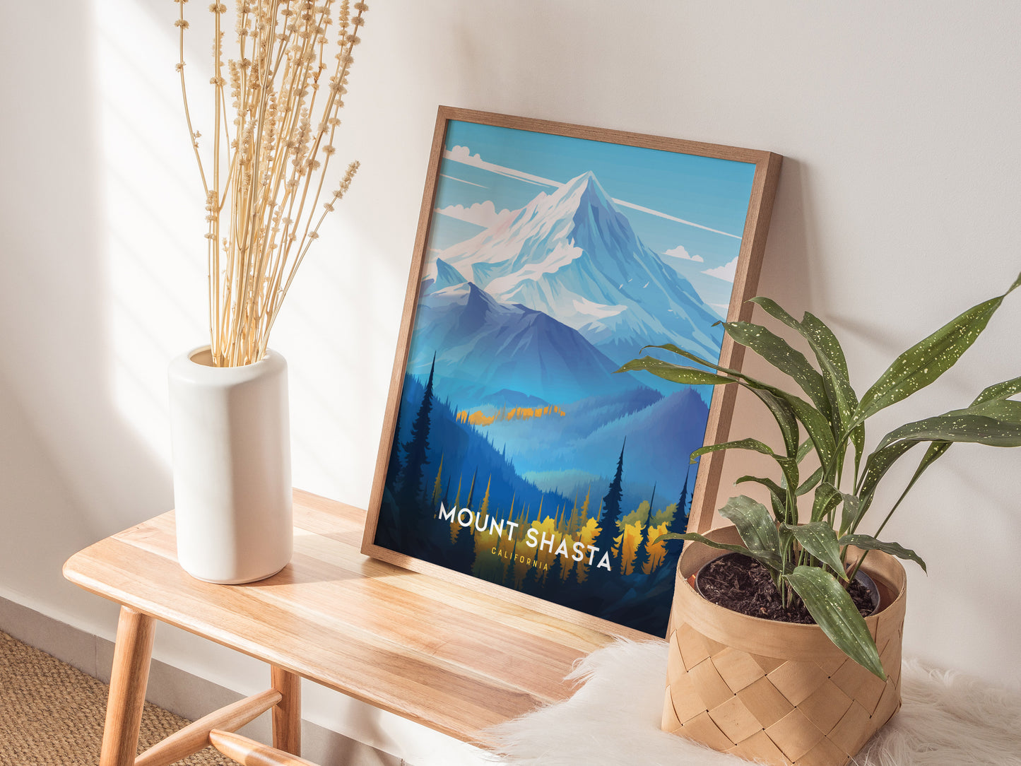 A framed Mount Shasta poster leans against a wall on a wooden bench, accompanied by a white vase with dried flowers and a potted plant in a woven basket.