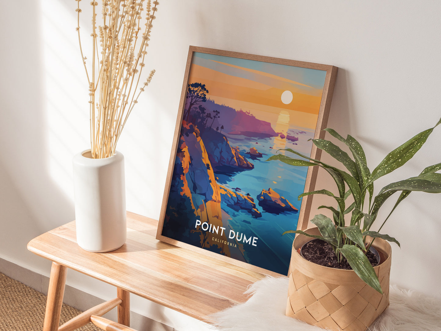 A framed Point Dume poster with vibrant coastal scenery rests on a wooden bench between a white vase with dried flowers and a potted green plant.