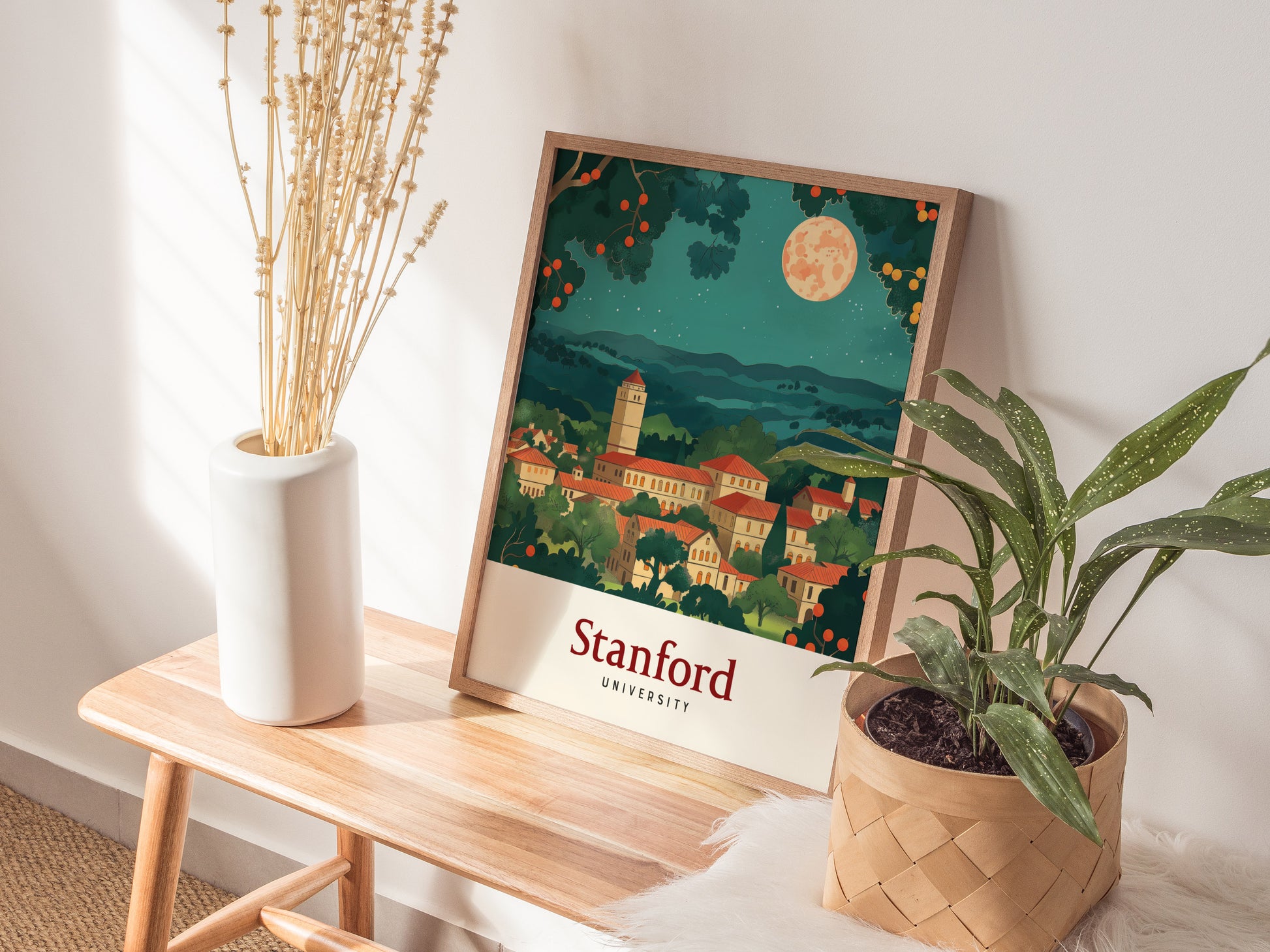 A framed poster of Stanford University rests on a wooden bench, surrounded by a white vase with dried flowers and a potted green plant. Sunlight casts soft shadows.