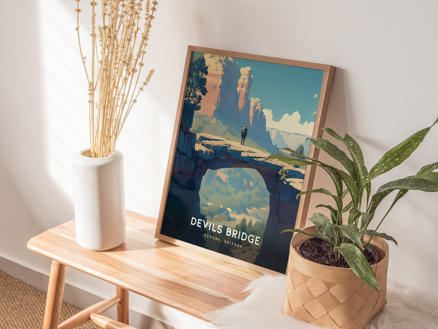 A framed Devils Bridge poster rests on a wooden bench beside a white vase with dried flowers and a potted green plant, creating a serene, natural aesthetic.