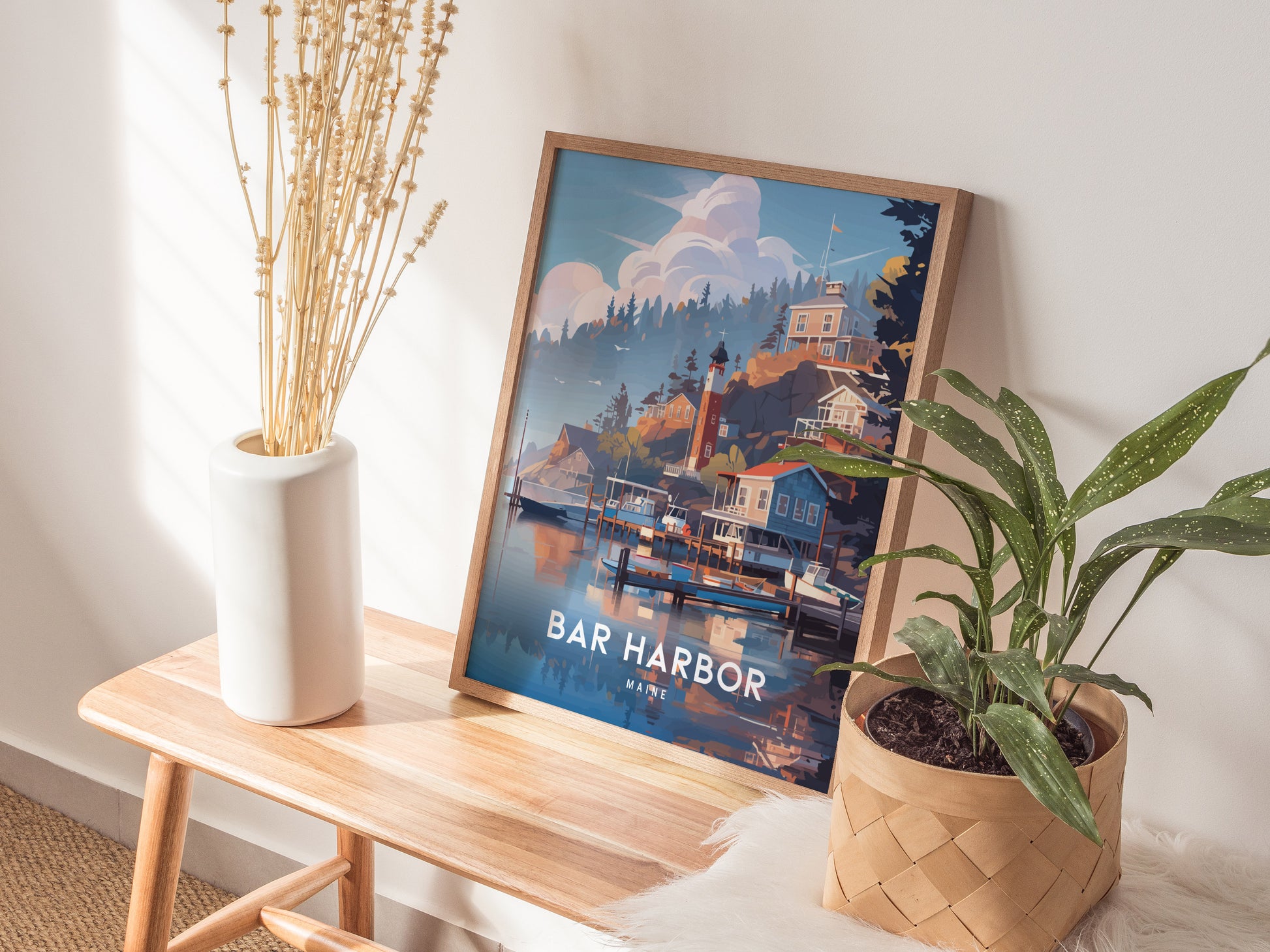 A framed Bar Harbor, Maine poster rests on a wooden bench beside a white vase with dried flowers and a green potted plant. Natural light illuminates the scene.