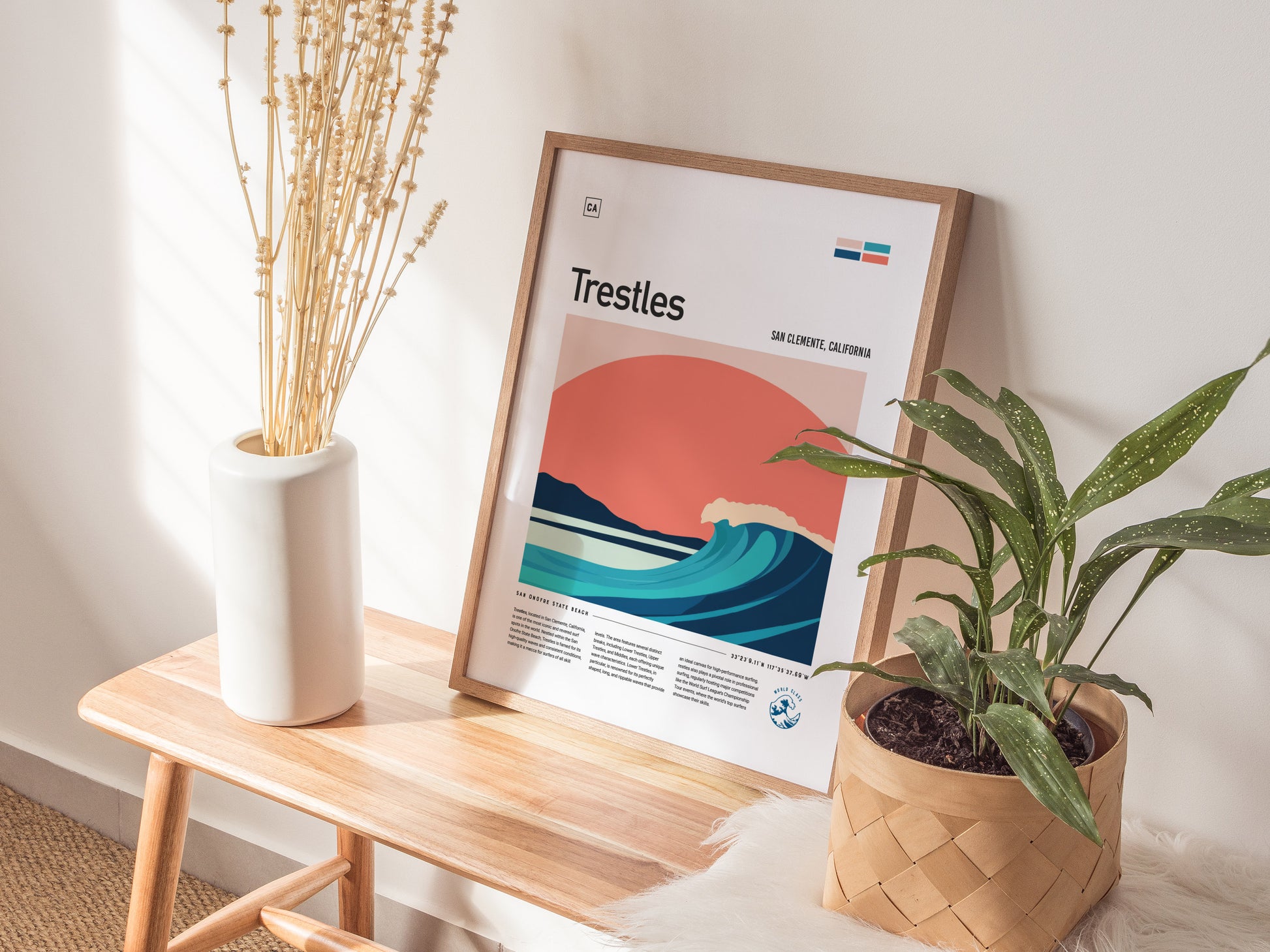 A framed Trestles surfer poster with vibrant wave art leans against a wall on a wooden bench, accompanied by a white vase with dried stems and a potted green plant.