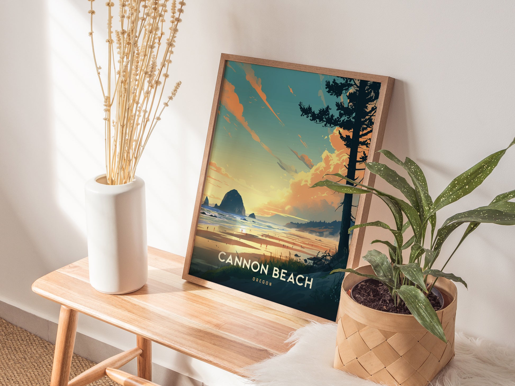 A Cannon Beach, Oregon poster with Haystack Rock is propped against a wall, next to dried flowers in a white vase and a potted plant on a wooden bench.