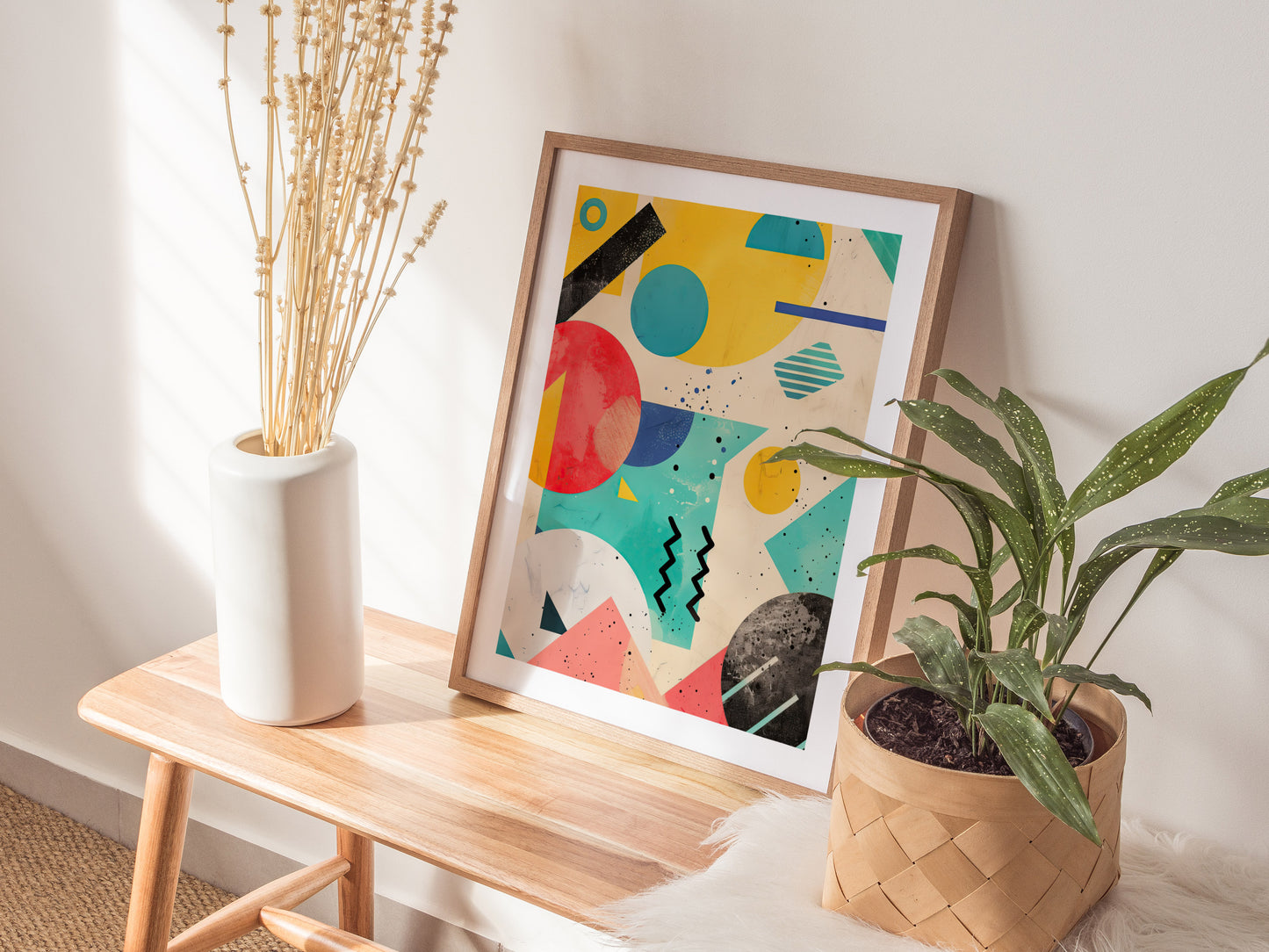 A colorful geometric art print with bold shapes is propped on a wooden bench, flanked by a white vase with dried flowers and a green potted plant.