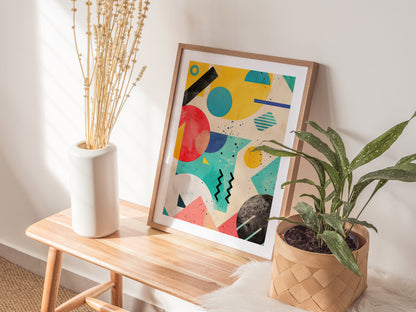 A colorful geometric art print with bold shapes is propped on a wooden bench, flanked by a white vase with dried flowers and a green potted plant.