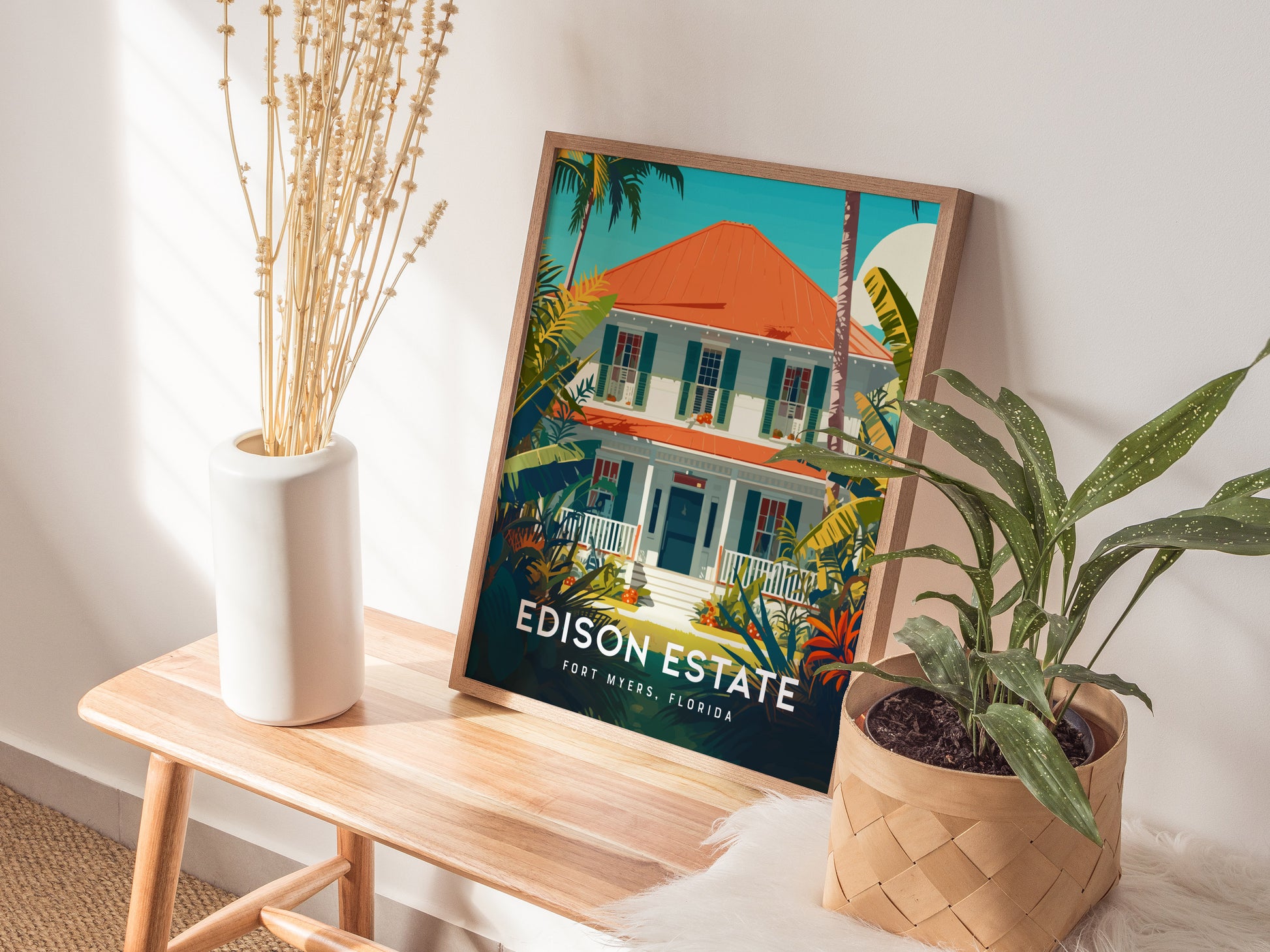 A framed poster titled Edison Estate, Fort Myers, Florida rests on a wooden bench, accompanied by a white vase with dried stems and a potted plant.