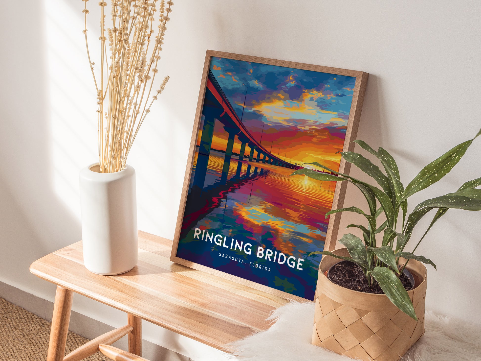 A framed poster of the Ringling Bridge in Sarasota, Florida, featuring a vibrant sunset, is displayed on a wooden bench. The setting includes a ceramic vase with dried flowers and a potted plant in a woven basket.