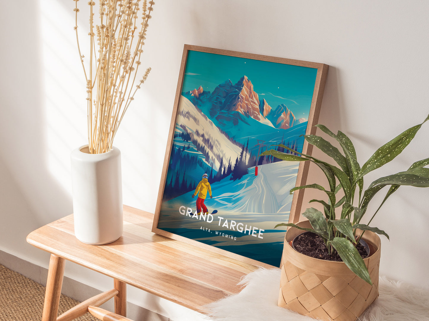A framed Grand Targhee Resort poster featuring snowy mountains and a snowboarder is displayed on a wooden bench beside a white vase with dried plants and a potted green plant.