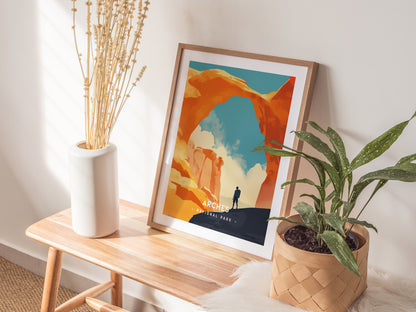 A framed minimalist poster of Arches National Park rests on a wooden bench, accompanied by a vase of dried flowers and a potted plant. Natural light illuminates the scene.