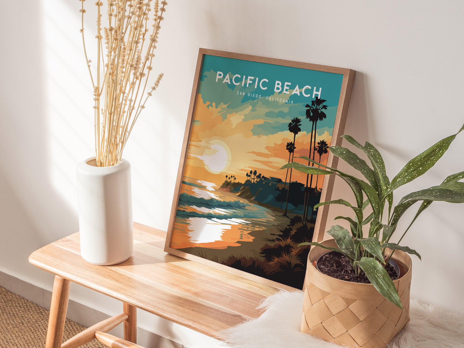 A framed Pacific Beach San Diego poster sits on a wooden bench, showcasing a vibrant sunset with palm trees and ocean waves. It is surrounded by a white vase with dried plants and a green potted plant, creating a serene coastal vibe.
